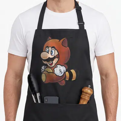 Tanooki holiday apron - Cartoon Tanooki Mario illustration in orange and tan with striped tail on black background — shown on a black cotton canvas apron against a white background