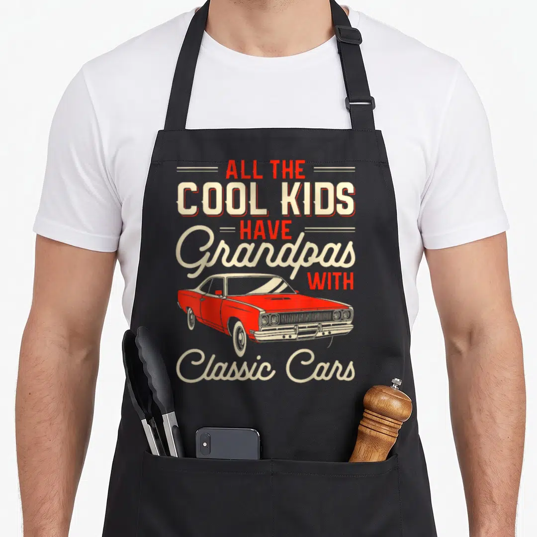 Grandpas car apron - Red classic car illustration surrounded by stacked red and beige typography reading ALL THE COOL — shown on a black cotton canvas apron against a white background