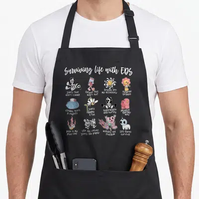 Surviving eds awareness apron - Twelve cartoon icons with captions about EDS symptoms like braces and fatigue — shown on a black cotton canvas apron against a white background