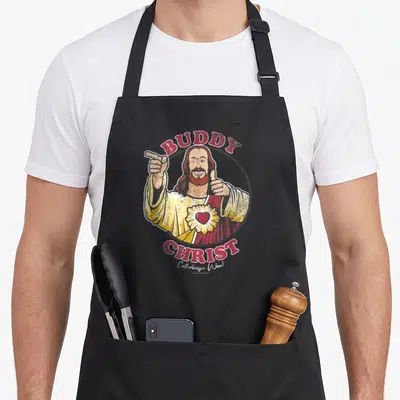 Jay buddy aprons - Distressed Buddy Christ portrait with Catholicism Wow! text in red and yellow — shown on a black cotton canvas apron against a white background
