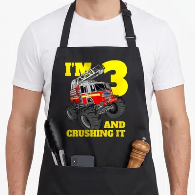 Monster fire truck apron - Red monster truck fire engine with yellow number 3 and text I'M 3 AND CRUSHING IT — shown on a black cotton canvas apron against a white background