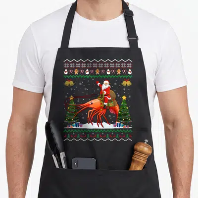 Shrimp ugly christmas apron - Santa riding giant red shrimp between Christmas trees with holiday icon borders — shown on a black cotton canvas apron against a white background