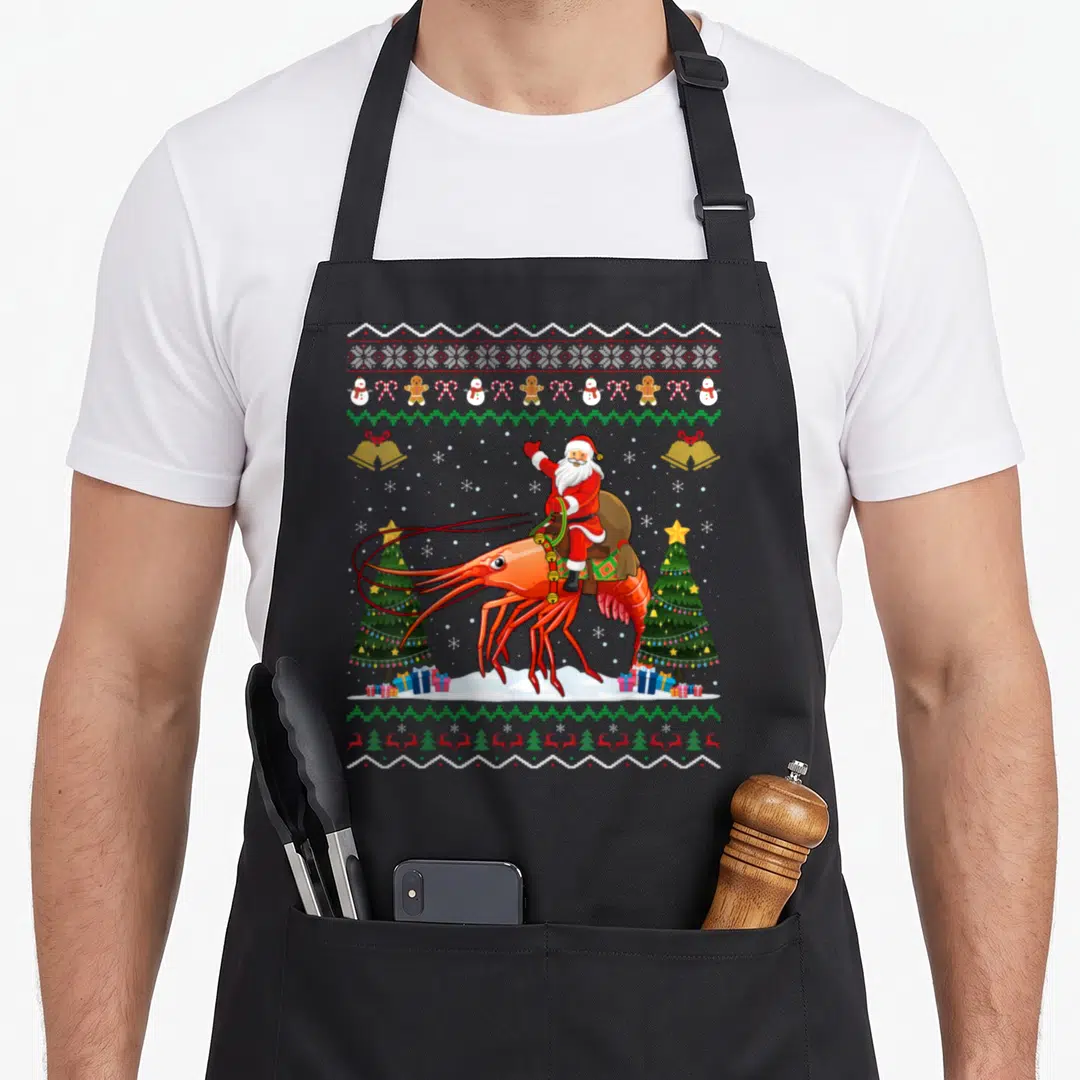 Shrimp ugly christmas apron - Santa riding giant red shrimp between Christmas trees with holiday icon borders — shown on a black cotton canvas apron against a white background