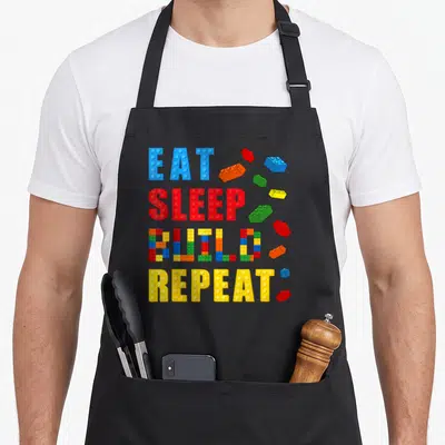 Eat block & apron - EAT SLEEP BUILD REPEAT text in block letters with scattered blue red yellow green orange bricks — shown on a black cotton canvas apron against a white background