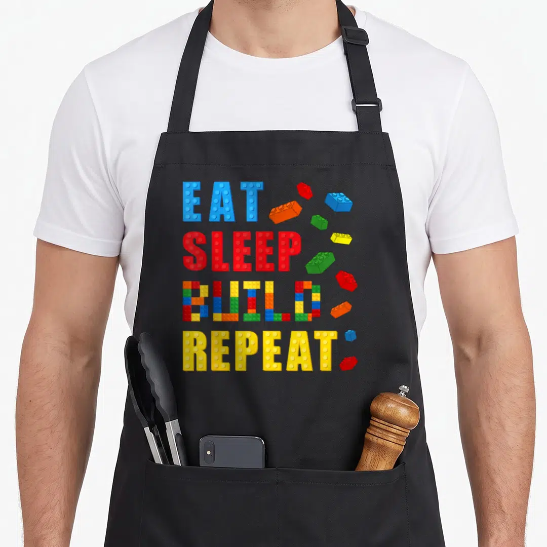 Eat block & apron - EAT SLEEP BUILD REPEAT text in block letters with scattered blue red yellow green orange bricks — shown on a black cotton canvas apron against a white background