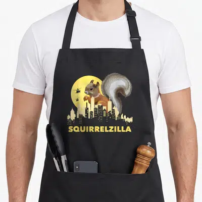 Squirrelzilla animal apron - Giant brown squirrel behind yellow city skyline with two helicopters and SQUIRRELZILLA text — shown on a black cotton canvas apron against a white background