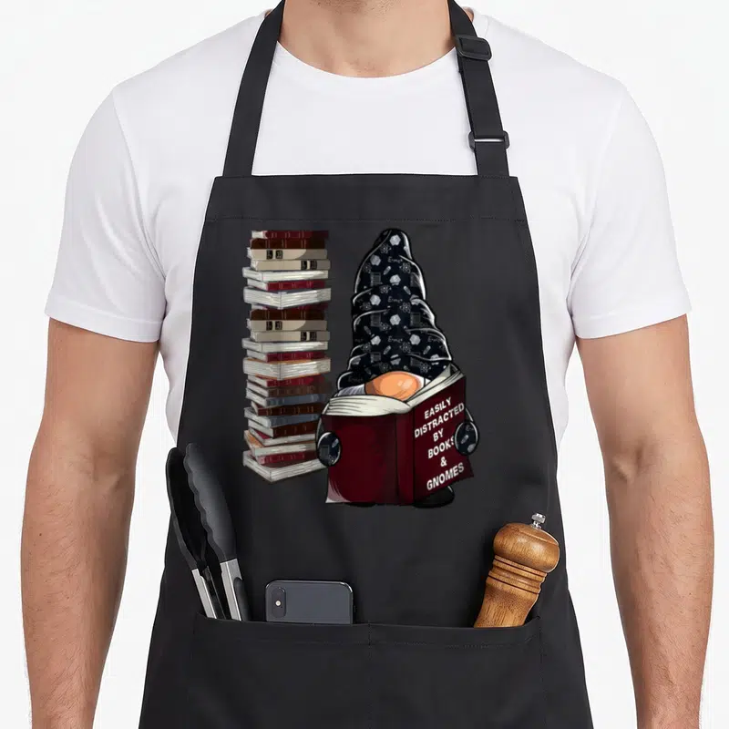 Distracted lover easily apron - Cartoon gnome holding book with stack of books and science doodles on hat — shown on a black cotton canvas apron against a white background