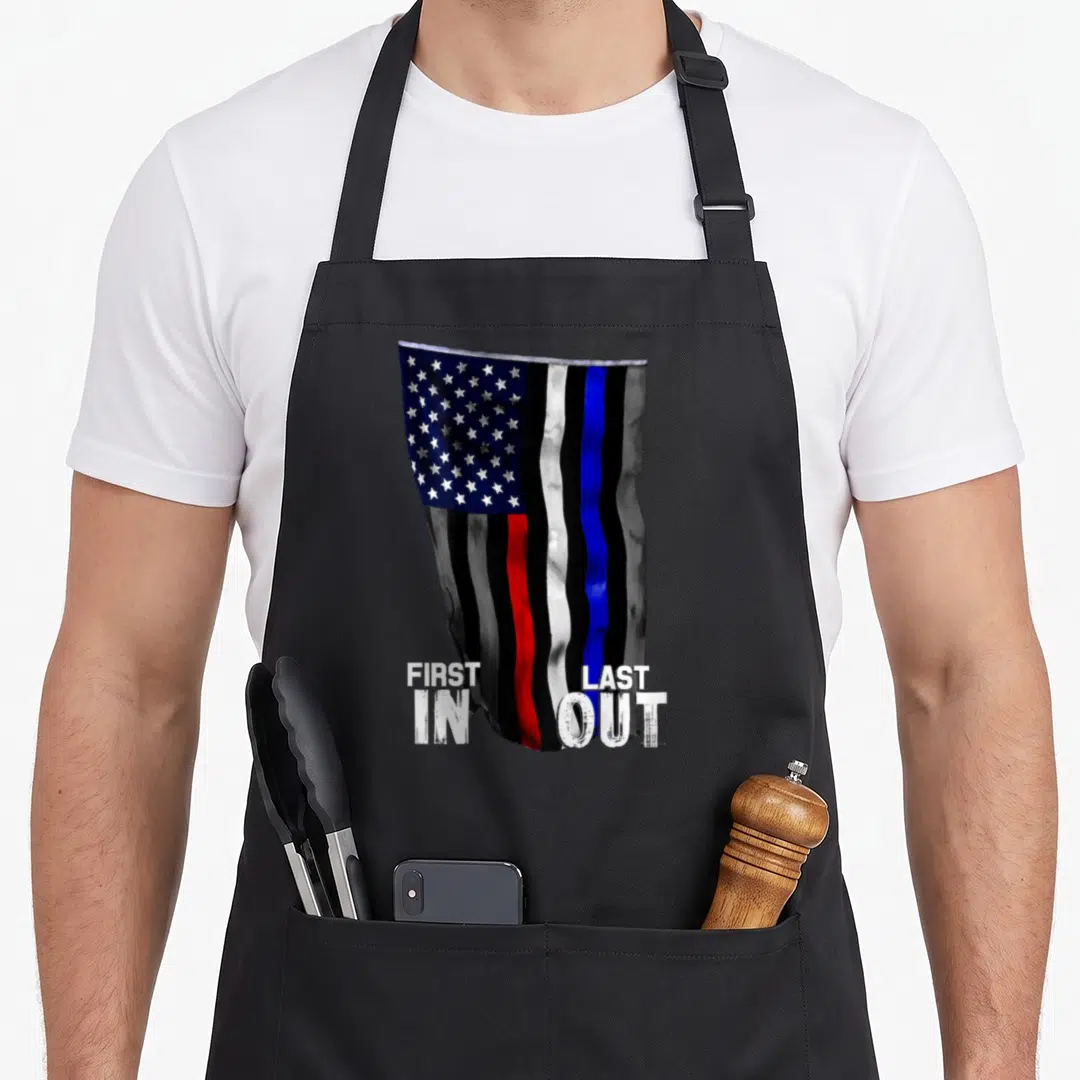 Fire american flag apron - Waving US flag with red white blue lines and FIRST IN LAST OUT text — shown on a black cotton canvas apron against a white background