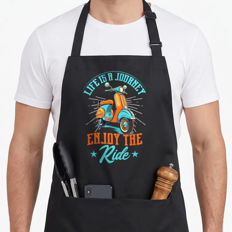 Journey scooter apron - Retro orange and blue scooter with LIFE IS A JOURNEY ENJOY THE Ride text and two stars — shown on a black cotton canvas apron against a white background