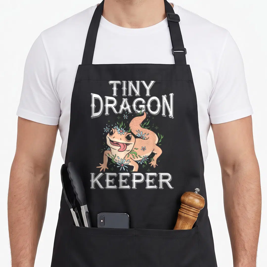 Tiny leopard apron - Cartoon gecko with TINY DRAGON KEEPER text surrounded by blue flowers and green leaves — shown on a black cotton canvas apron against a white background
