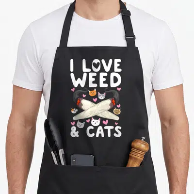 Cannabis cat apron - Crossed joints surrounded by cat heads and pink hearts with I LOVE WEED & CATS text — shown on a black cotton canvas apron against a white background