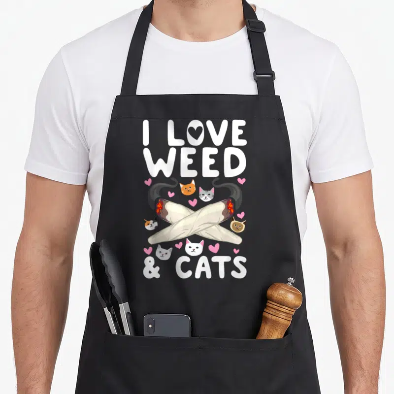 Cannabis cat apron - Crossed joints surrounded by cat heads and pink hearts with I LOVE WEED & CATS text — shown on a black cotton canvas apron against a white background