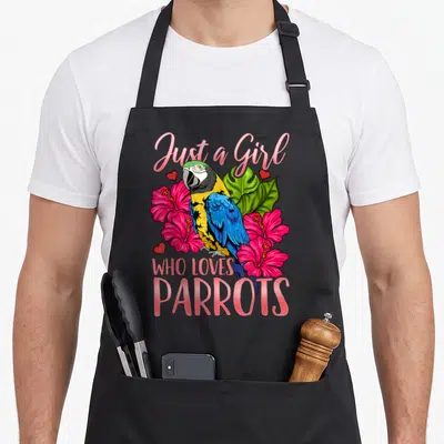 Parrot watching bird apron - Cartoon parrot with pink hibiscus flowers and text Just a Girl Who Loves Parrots — shown on a black cotton canvas apron against a white background