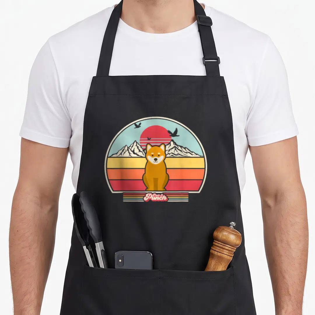 Shiba dog husband apron - Retro shiba inu sitting before striped sun and mountains with Pack A Punch banner below — shown on a black cotton canvas apron against a white background