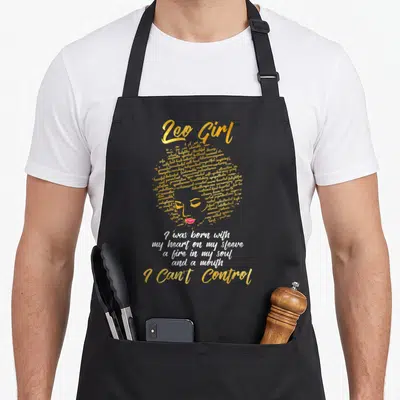 Retro leo birthday apron - Gold typography woman face with pink lips and white quote I was born with my heart on my — shown on a black cotton canvas apron against a white background