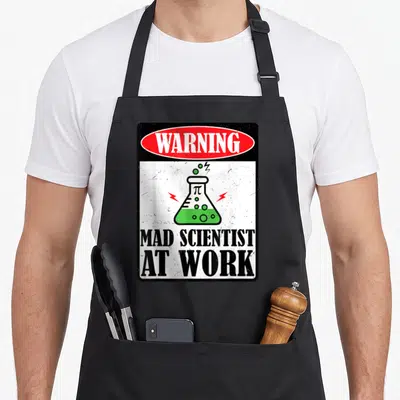 Mad teacher apron - Red warning oval, green flask, black mad scientist text on white background — shown on a black cotton canvas apron against a white background