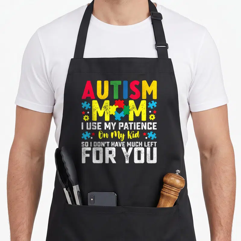 Autism use of apron - Colorful puzzle piece heart with AUTISM MOM text and scattered stars flowers — shown on a black cotton canvas apron against a white background