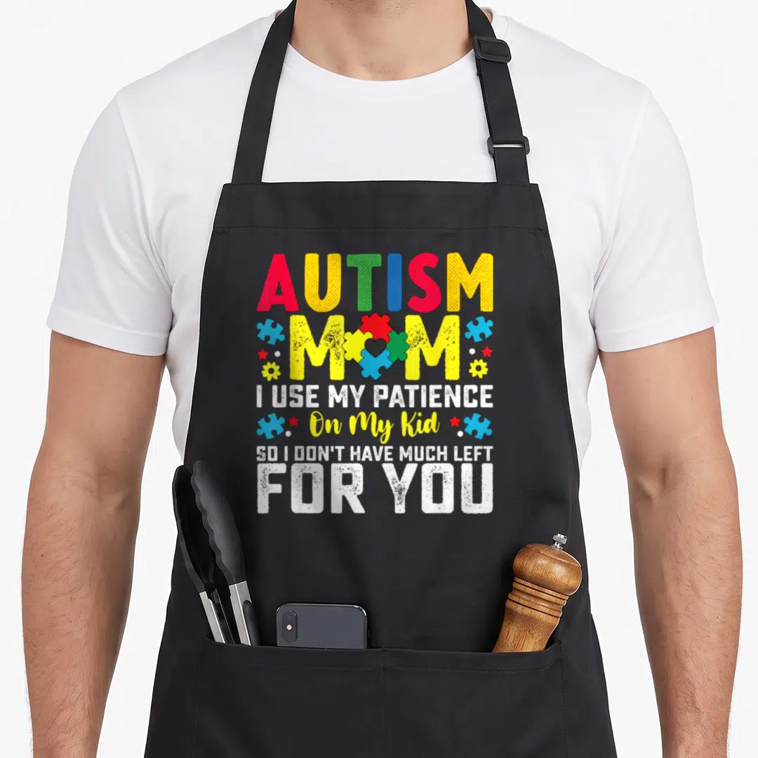 Autism use of apron - Colorful puzzle piece heart with AUTISM MOM text and scattered stars flowers — shown on a black cotton canvas apron against a white background