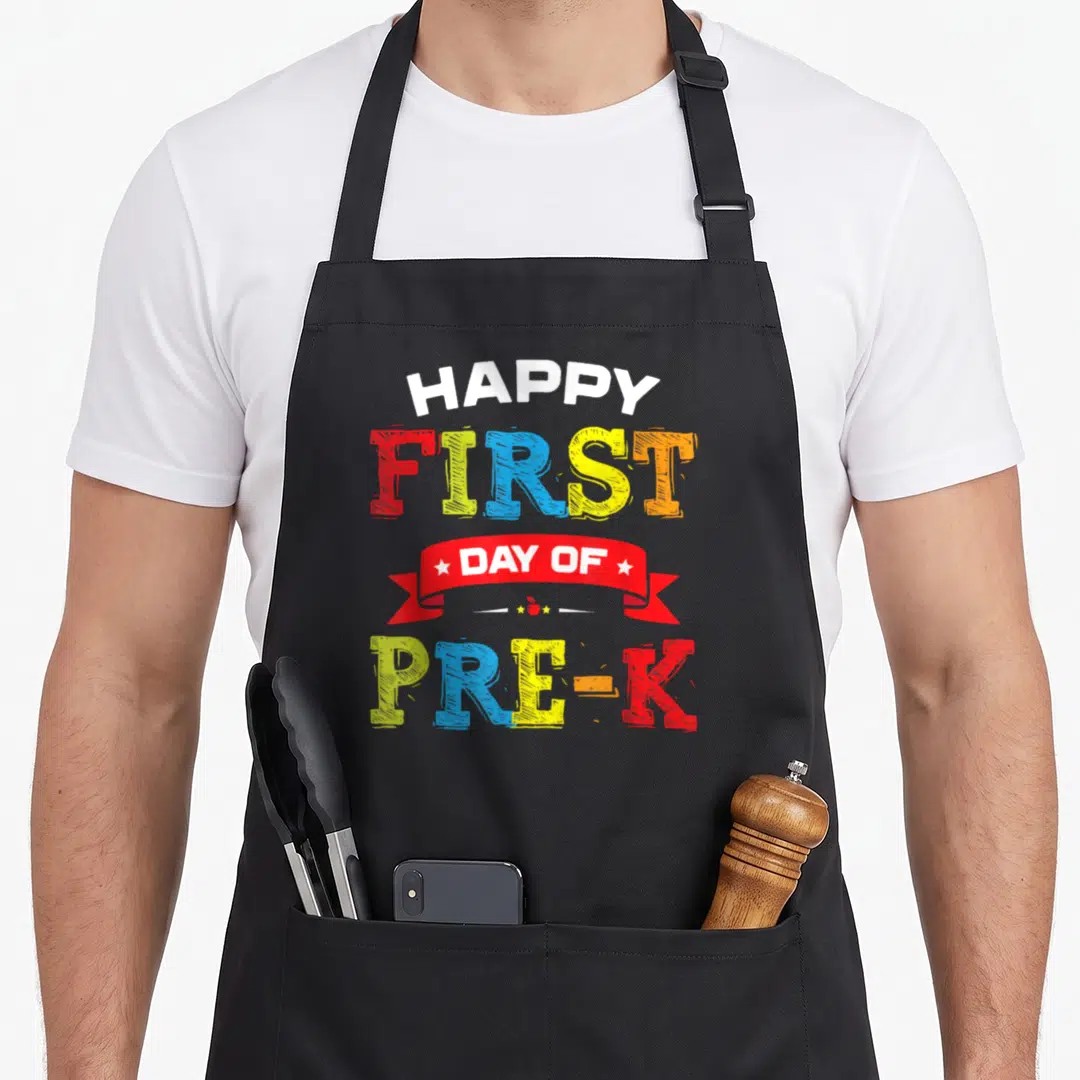 Pre teacher preschool apron - Stacked text Happy First Day Of Pre-K in red yellow blue green white with ribbon and apple — shown on a black cotton canvas apron against a white background