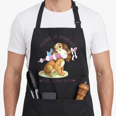 Funny dog lady and the tramp apron - Brown cartoon dog holding pink knitting needles on grass with arched pink text — shown on a black cotton canvas apron against a white background