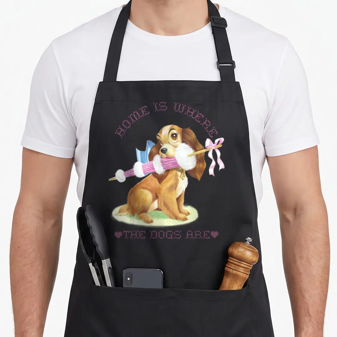 Funny dog lady and the tramp apron - Brown cartoon dog holding pink knitting needles on grass with arched pink text — shown on a black cotton canvas apron against a white background