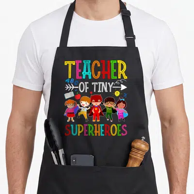 Prek kindergarten teacher apron - Five cartoon children in superhero costumes flanked by book apple lightbulb percent — shown on a black cotton canvas apron against a white background