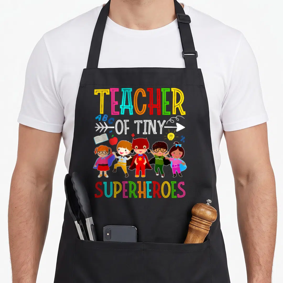 Prek kindergarten teacher apron - Five cartoon children in superhero costumes flanked by book apple lightbulb percent — shown on a black cotton canvas apron against a white background