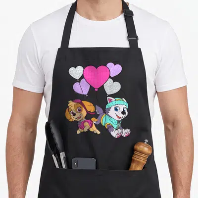 Skye balloon apron - Two cartoon puppies holding five heart balloons in pink purple and blue — shown on a black cotton canvas apron against a white background