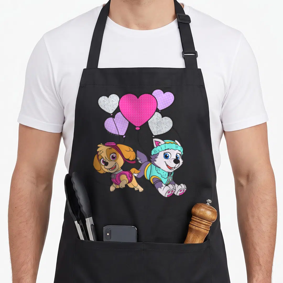 Skye balloon apron - Two cartoon puppies holding five heart balloons in pink purple and blue — shown on a black cotton canvas apron against a white background