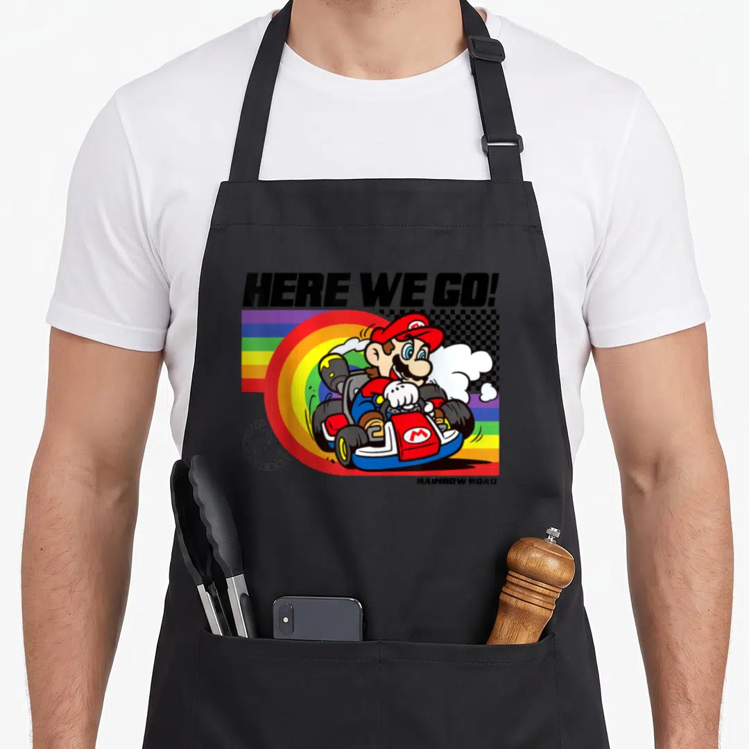 Pride road apron - Mario karting over rainbow graphic with HERE WE GO! and RAINBOW ROAD text — shown on a black cotton canvas apron against a white background