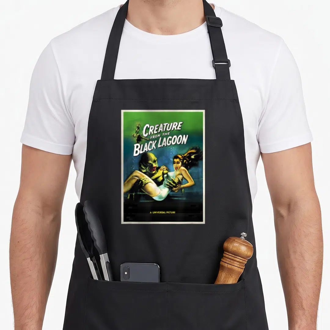 Creature ogre apron - Retro Creature from the Black Lagoon poster art in green and blue showing a creature holding — shown on a black cotton canvas apron against a white background