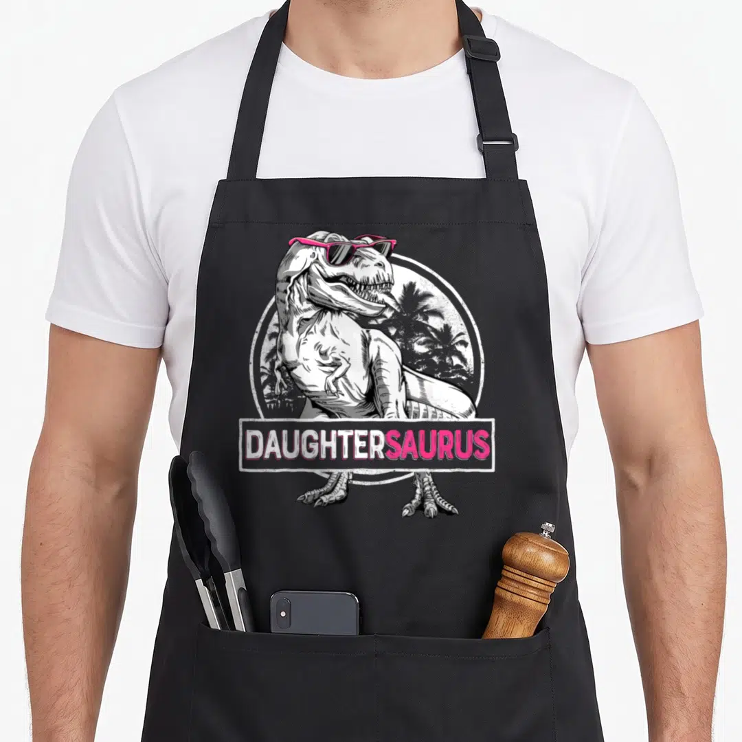 Daughtersaurus mom daughter aprons - T-Rex in pink sunglasses with DAUGHTERSAURUS banner and palm tree background — shown on a black cotton canvas apron against a white background