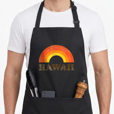 Souvenir rainbow apron - Distressed retro rainbow arch above brown HAWAII text — shown on a black cotton canvas apron against a white background
