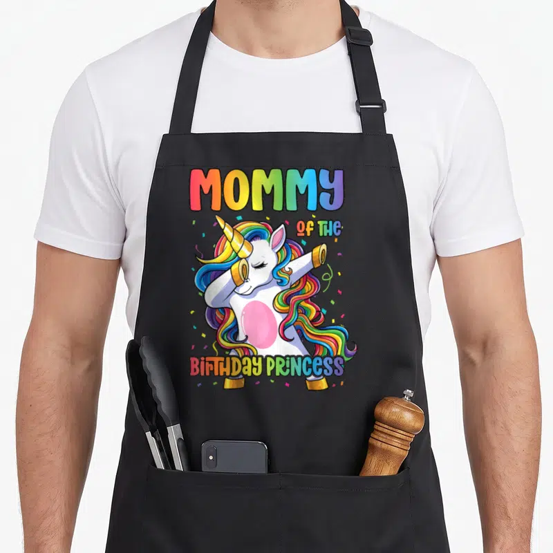Dabbing princess apron - Rainbow unicorn dabbing with MOMMY OF THE BIRTHDAY PRINCESS text and confetti — shown on a black cotton canvas apron against a white background