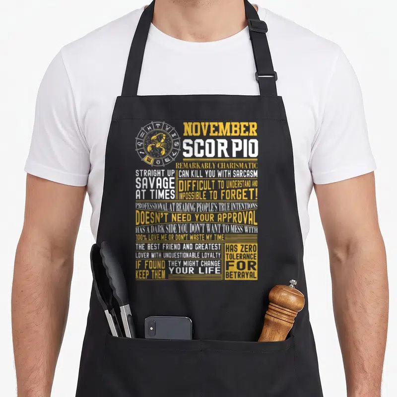 November scorpio girl apron - Zodiac wheel with scorpion and stacked yellow white text listing Scorpio personality — shown on a black cotton canvas apron against a white background