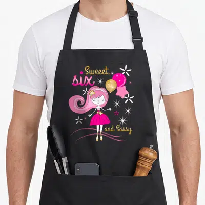Birthday sweet sassy apron - Cartoon girl holding pink and gold balloons with Sweeet SIX and Sassy text — shown on a black cotton canvas apron against a white background