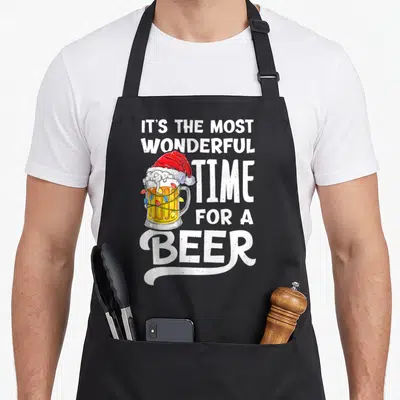 Most santa aprons - Beer mug wearing red Santa hat wrapped in colored lights beside white stacked text — shown on a black cotton canvas apron against a white background