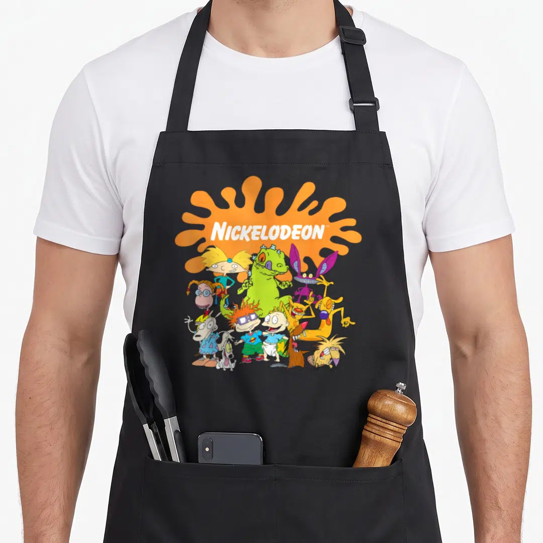 Funny shot character aprons - NICKELODEON text over orange splat with cartoon characters in green, blue, yellow, purple — shown on a black cotton canvas apron against a white background