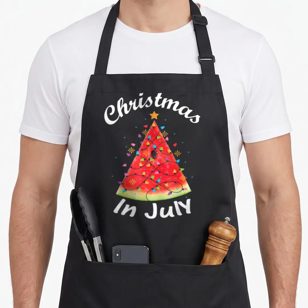 July christmas tree apron - Watermelon slice shaped like a Christmas tree wrapped in colorful lights with white text — shown on a black cotton canvas apron against a white background