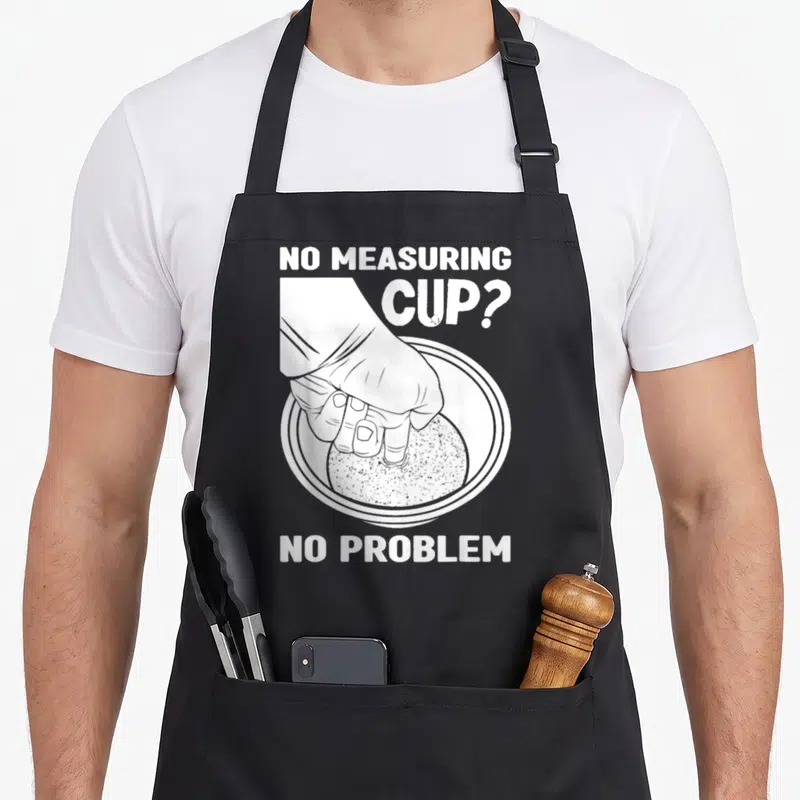 Filipino graphic problems apron - White hand pressing into rice bowl with text no measuring cup no problem — shown on a black cotton canvas apron against a white background