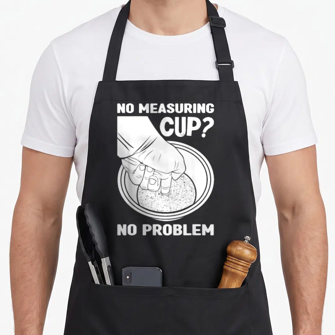Filipino graphic problems apron - White hand pressing into rice bowl with text no measuring cup no problem — shown on a black cotton canvas apron against a white background