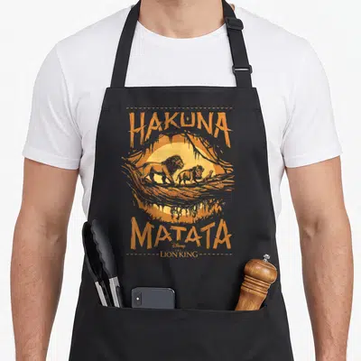 Live king of apron - Retro graphic of three lions walking before a sun with Hakuna Matata text in orange and yellow — shown on a black cotton canvas apron against a white background