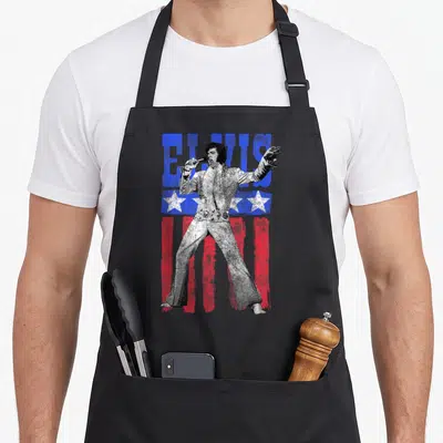 Elvis american flag apron - Distressed portrait of Elvis Presley singing with blue ELVIS text and red white flag — shown on a black cotton canvas apron against a white background