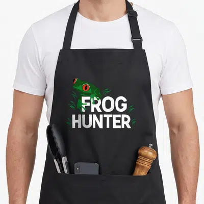 Frog special aprons - Green frog with red eyes over white FROG HUNTER text with grass tufts — shown on a black cotton canvas apron against a white background