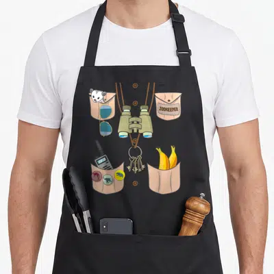 Keeper halloween apron - Cartoon zookeeper vest with binoculars, lemur, sunglasses, walkie-talkie, keys, and bananas — shown on a black cotton canvas apron against a white background