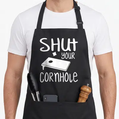 Shut game aprons - White SHUT your CORNHOLE text with bean bag and board illustration — shown on a black cotton canvas apron against a white background