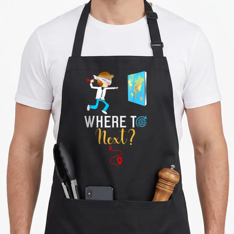 International traveler apron - Blindfolded man darting at map with WHERE TO Next text and red route pins — shown on a black cotton canvas apron against a white background