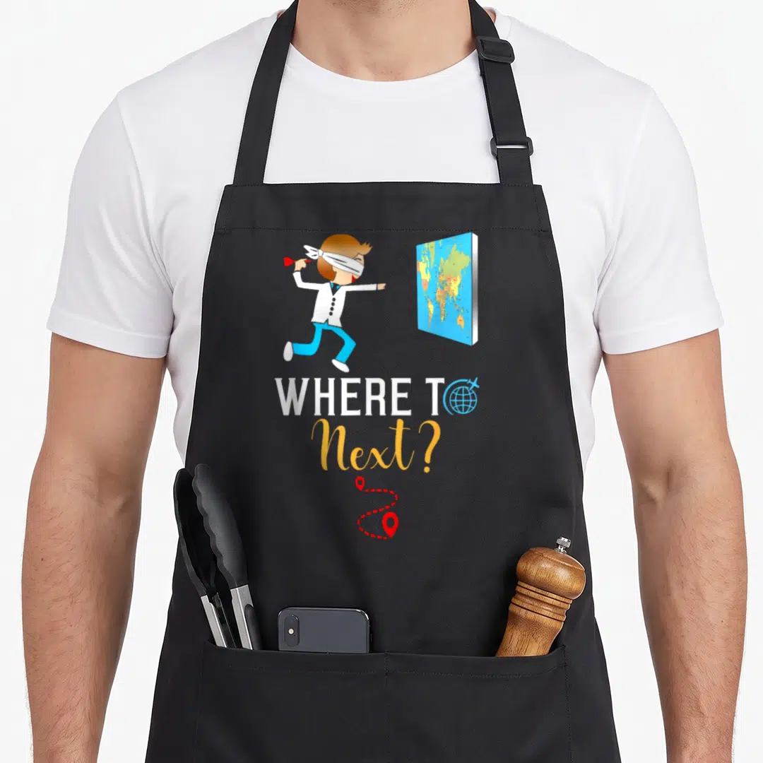 International traveler apron - Blindfolded man darting at map with WHERE TO Next text and red route pins — shown on a black cotton canvas apron against a white background