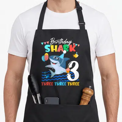 Baby birthday apron - Cartoon shark wearing party hat holding balloons and cupcake next to number 3 and THREE THREE — shown on a black cotton canvas apron against a white background
