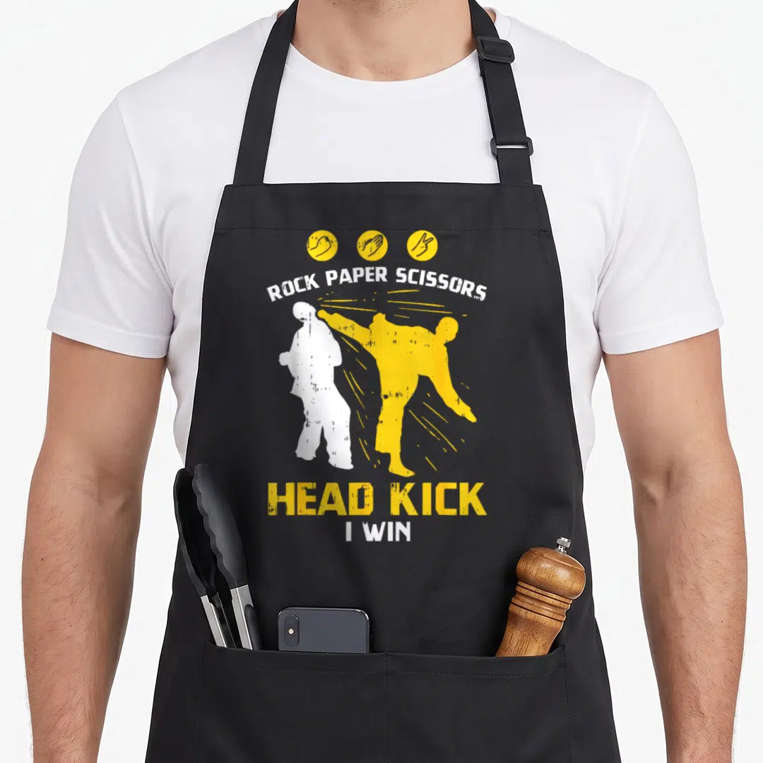 Headkick paper apron - Yellow and white martial artist kicking figure with Rock Paper Scissors text and hand icons — shown on a black cotton canvas apron against a white background